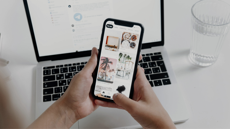 Mano sujetando un smartphone con un ordenador de fondo, ilustrando el proceso de crear productos digitales desde cualquier lugar